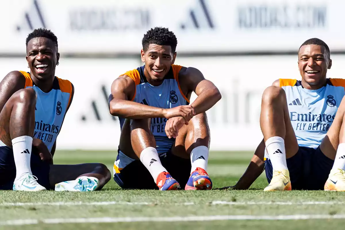 Three soccer players sitting on the grass during a training session, wearing Real Madrid training uniforms and smiling.