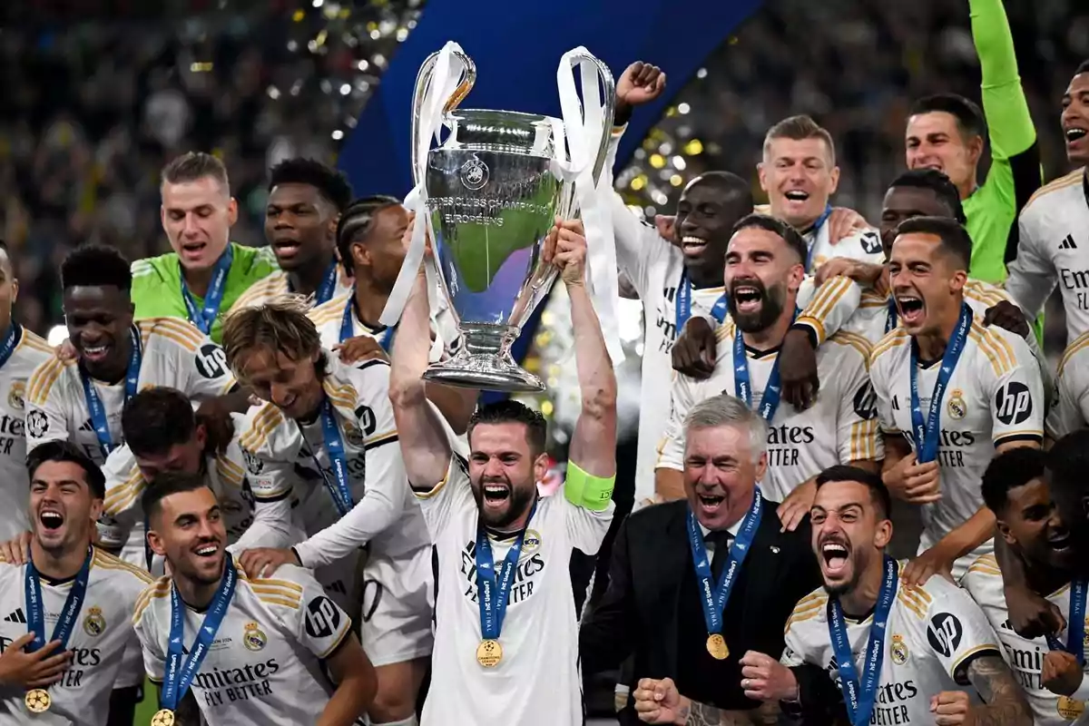 Jugadores de un equipo de fútbol celebran con un trofeo grande y plateado, todos visten uniformes blancos y medallas azules. Jugadores de un equipo de fútbol celebran con un trofeo grande y plateado, todos visten uniformes blancos y medallas azules.