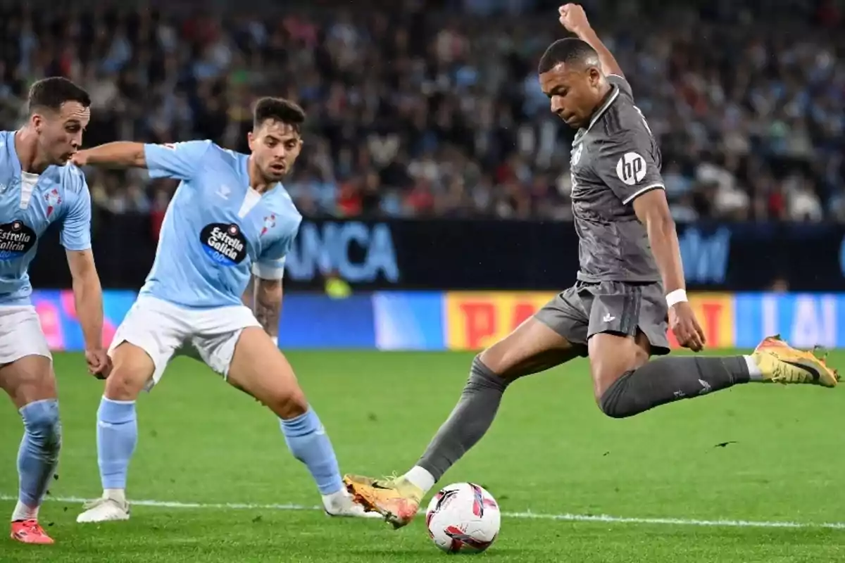 A soccer player in a gray uniform kicks the ball while two players in light blue uniforms watch him in a stadium full of spectators. A soccer player in a gray uniform kicks the ball while two players in light blue uniforms watch him in a stadium full of spectators.