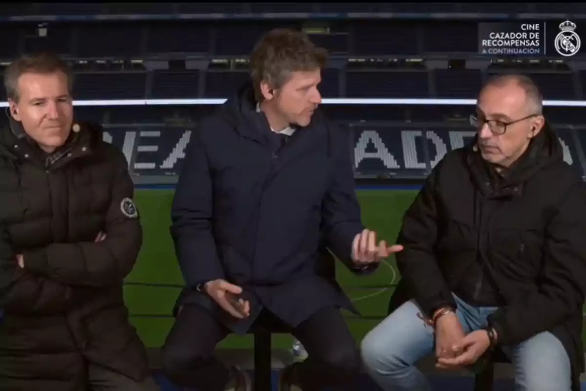 Three men sitting in a television studio with a football stadium in the background, one of them gesturing while the other two listen alertly.