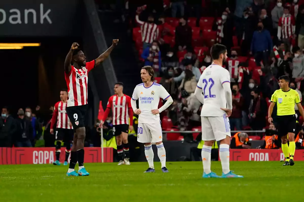 Soccer players on the field during a match, one of them raises their arms in celebration while others watch, with spectators in the stands. Soccer players on the field during a match, one of them raises their arms in celebration while others watch, with spectators in the stands.