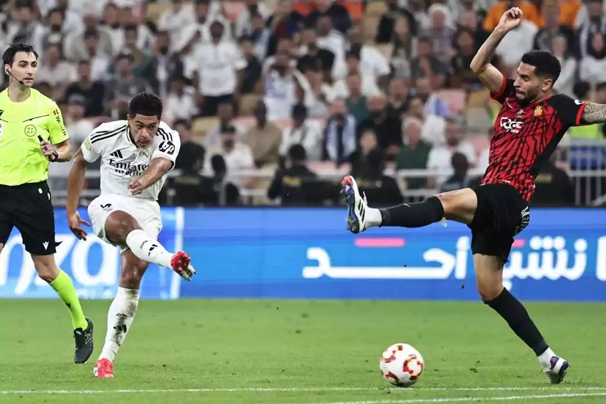 Um jogador de futebol de uniforme branco chuta a bola enquanto um adversário de uniforme vermelho e preto tenta bloquear a bola em um estádio lotado de espectadores.