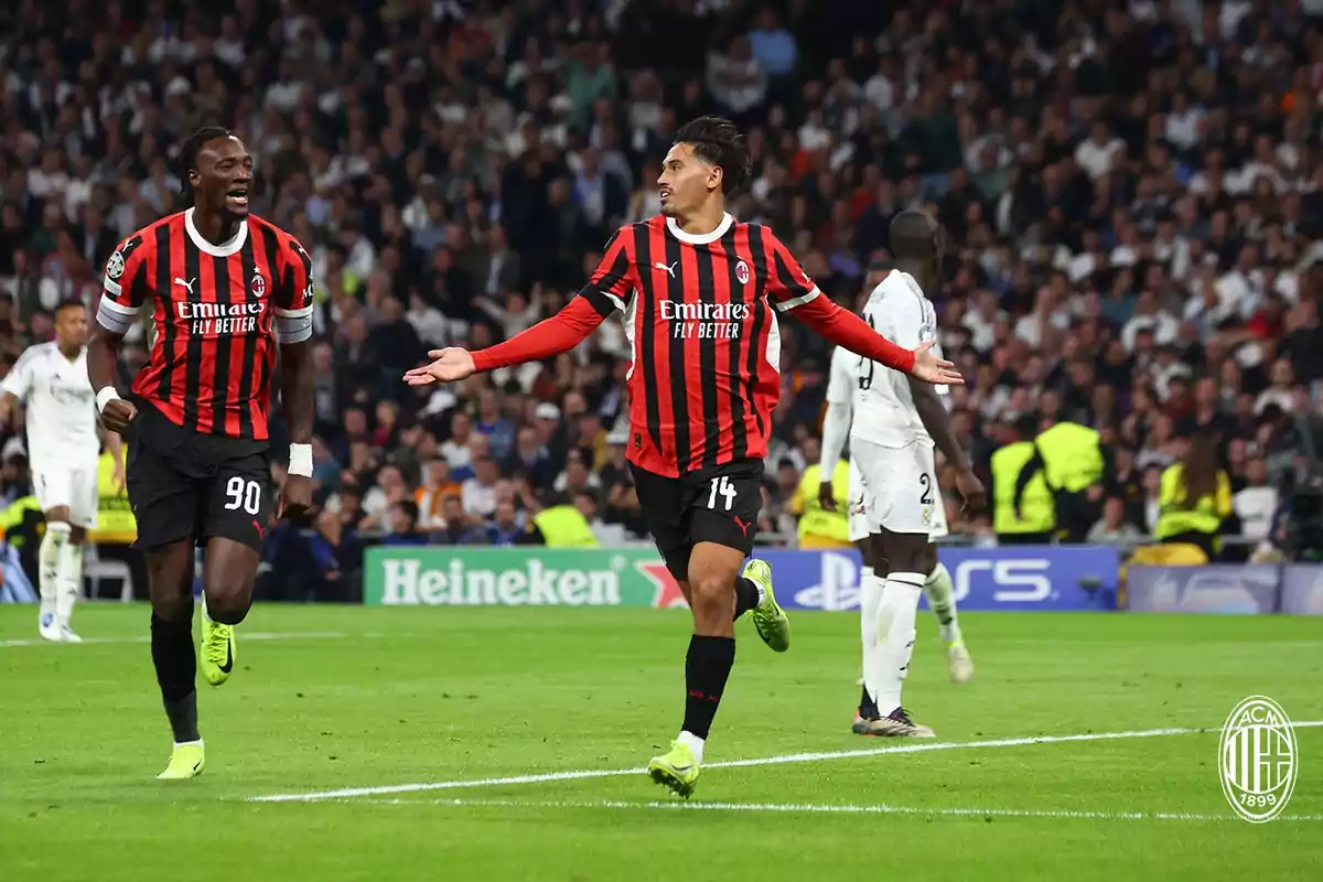 Two AC Milan players celebrate a goal in a packed stadium during a soccer match, while a player from the opposing team walks away with his back turned. Two AC Milan players celebrate a goal in a packed stadium during a soccer match, while a player from the opposing team walks away with his back turned.