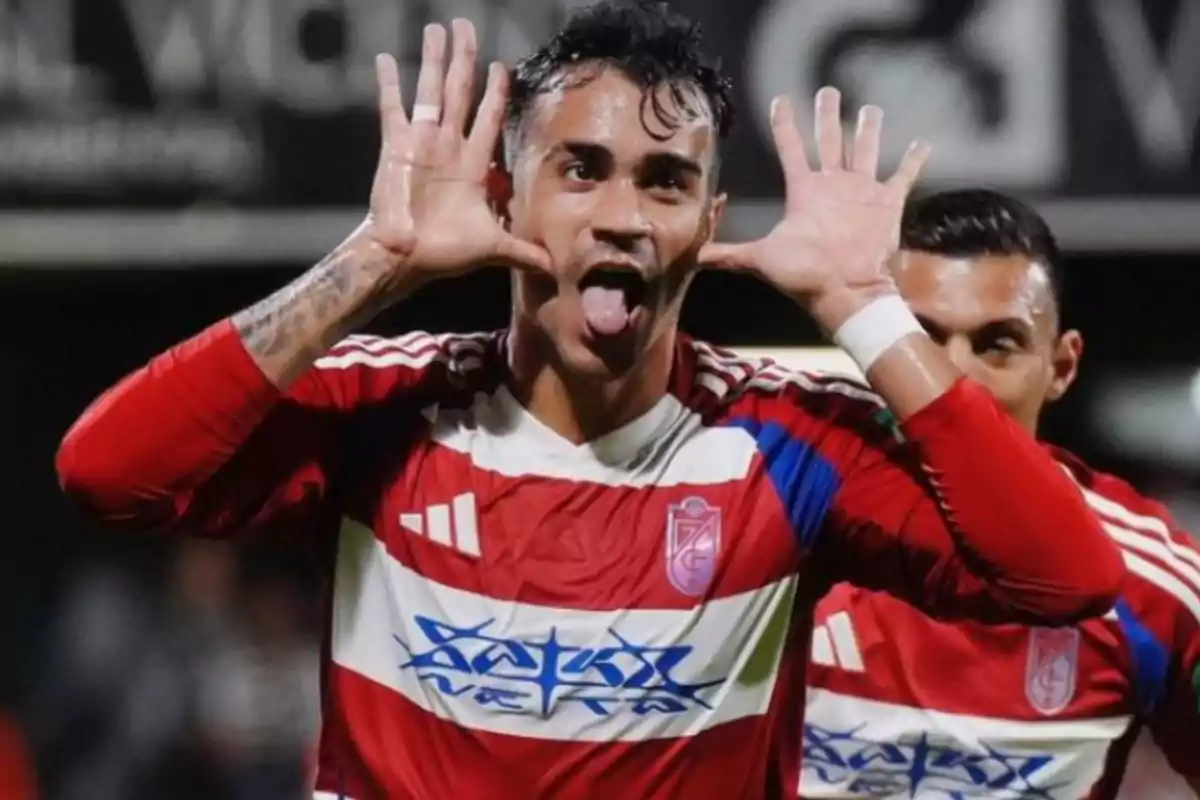 Soccer player in a red and white uniform celebrating a goal by making a funny gesture with his hands and sticking his tongue out Soccer player in a red and white uniform celebrating a goal by making a funny gesture with his hands and sticking his tongue out