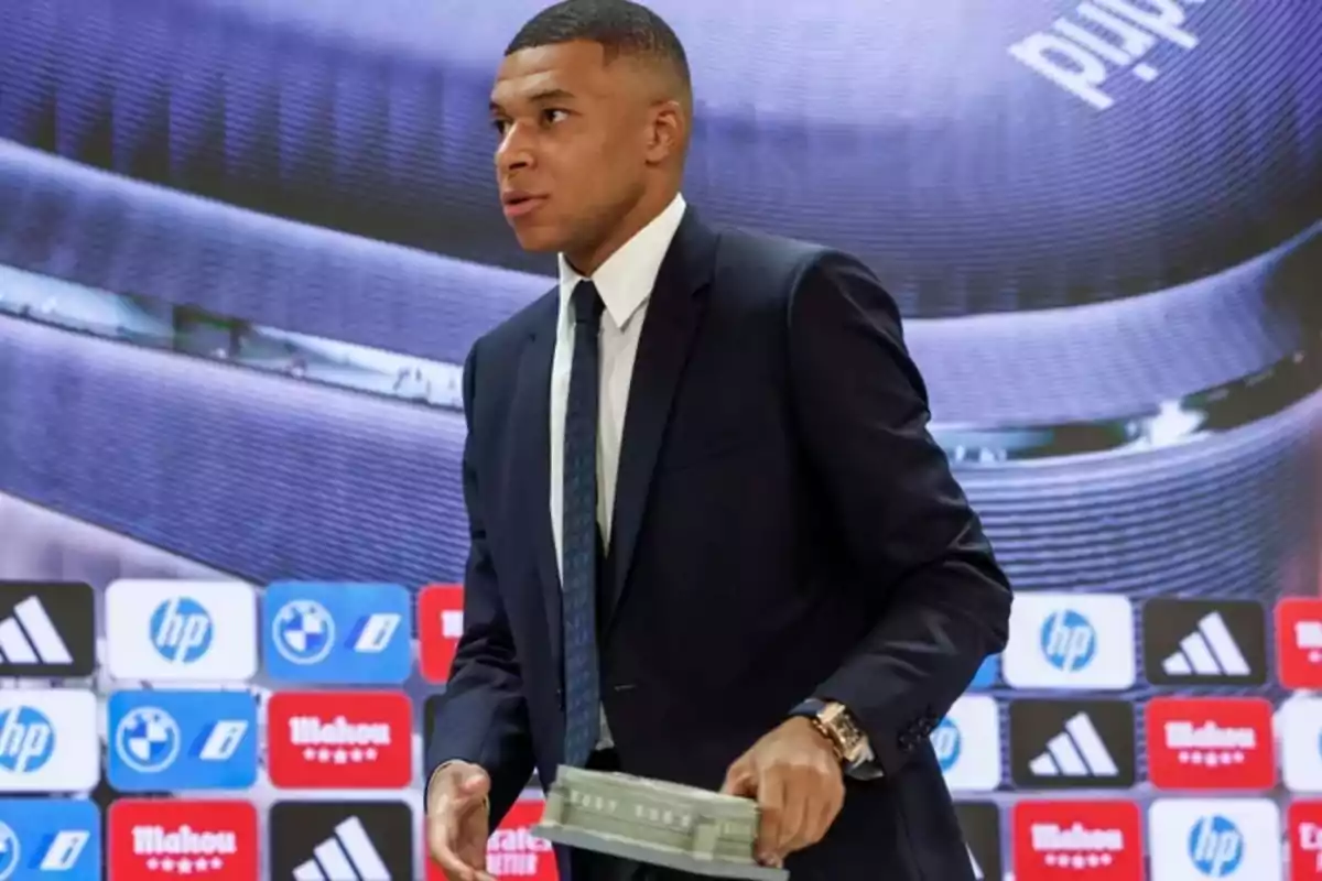 A man in a dark suit and tie holds a rectangular object in front of a background with brand logos. A man in a dark suit and tie holds a rectangular object in front of a background with brand logos.