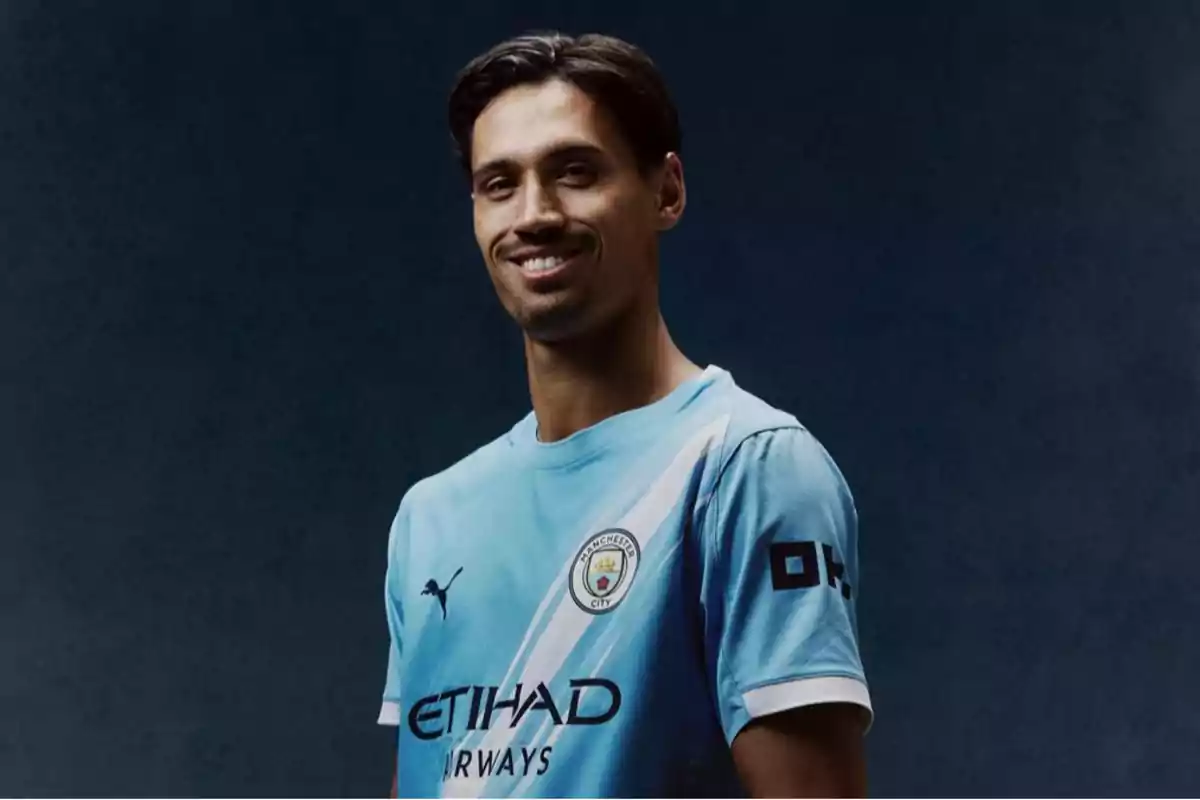 Jugador de fútbol con camiseta celeste del Manchester City posando y sonriendo frente a un fondo oscuro Jugador de fútbol con camiseta celeste del Manchester City posando y sonriendo frente a un fondo oscuro