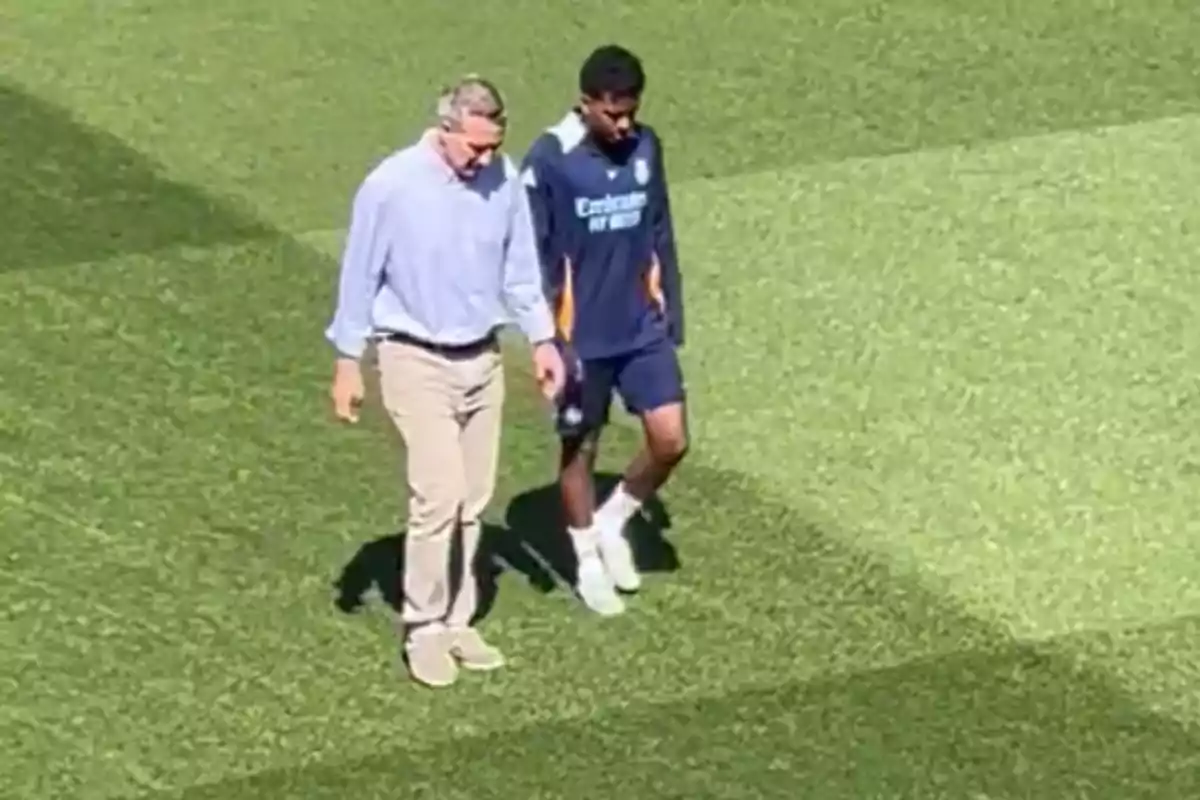 Dos personas caminando sobre el césped de un campo de fútbol, una vestida de manera casual y la otra con ropa deportiva. Dos personas caminando sobre el césped de un campo de fútbol, una vestida de manera casual y la otra con ropa deportiva.