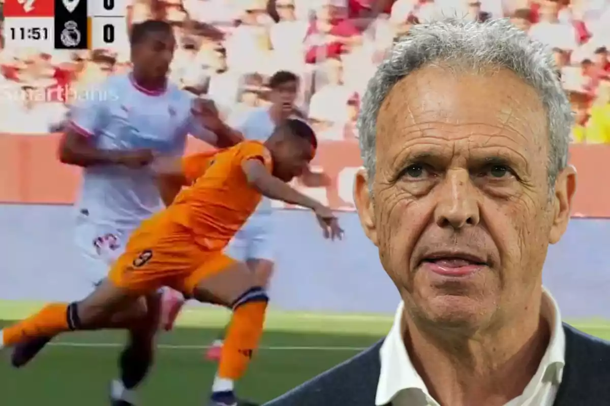 Un jugador de fútbol en uniforme naranja cae al suelo durante un partido mientras otro jugador con uniforme blanco está cerca y un hombre mayor aparece en primer plano.
