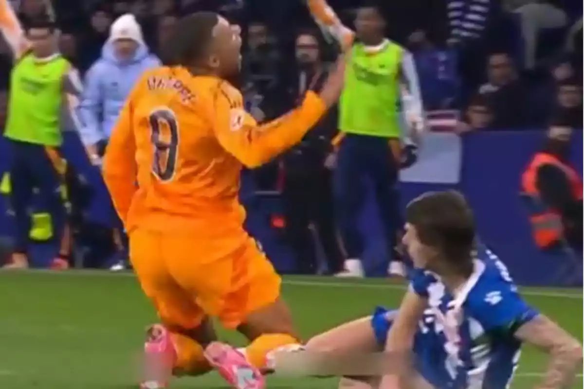 A player in an orange uniform falls to the ground while another player in a blue and white uniform sits on the grass during a match.