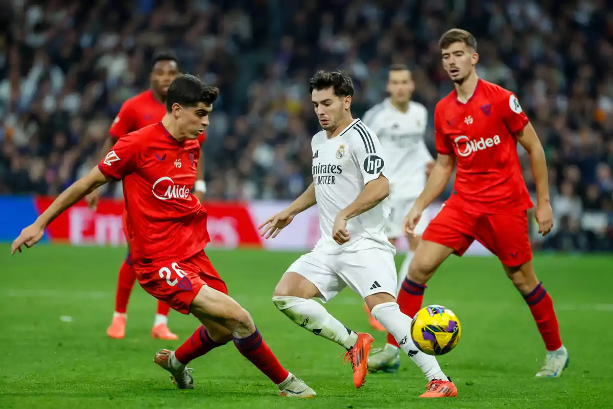 Jugadores de fútbol en acción durante un partido, uno con uniforme blanco y otros con uniforme rojo, en un estadio lleno de espectadores.