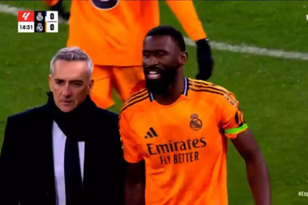 A player in an orange Real Madrid uniform next to a man with a coat and scarf on the playing field.