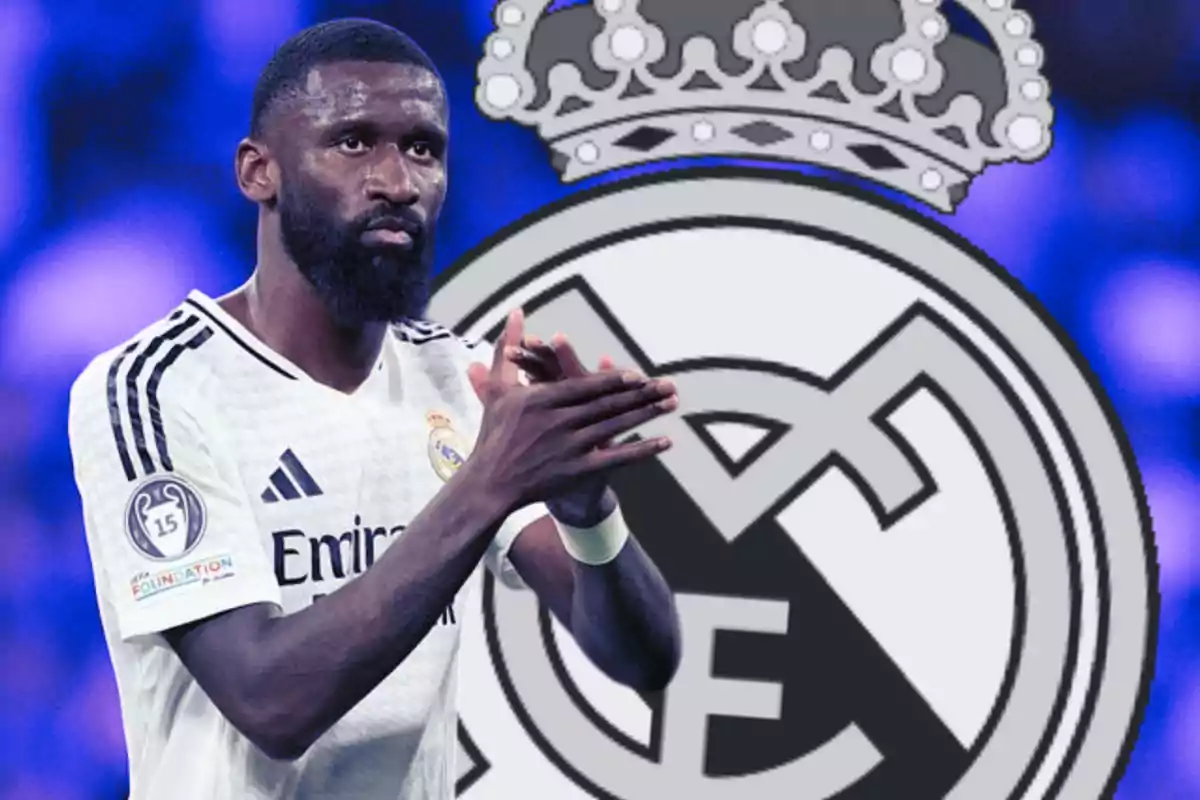 Un jugador de fútbol con uniforme blanco aplaude frente a un escudo grande del Real Madrid.