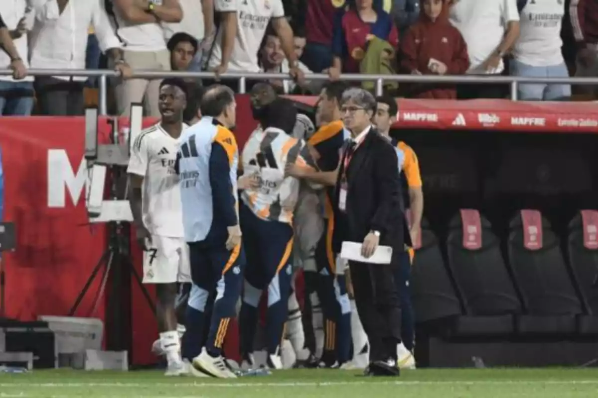 A group of people in a soccer stadium, some in sports uniforms and others in casual clothing, interact near the sideline while the audience watches from the stands.