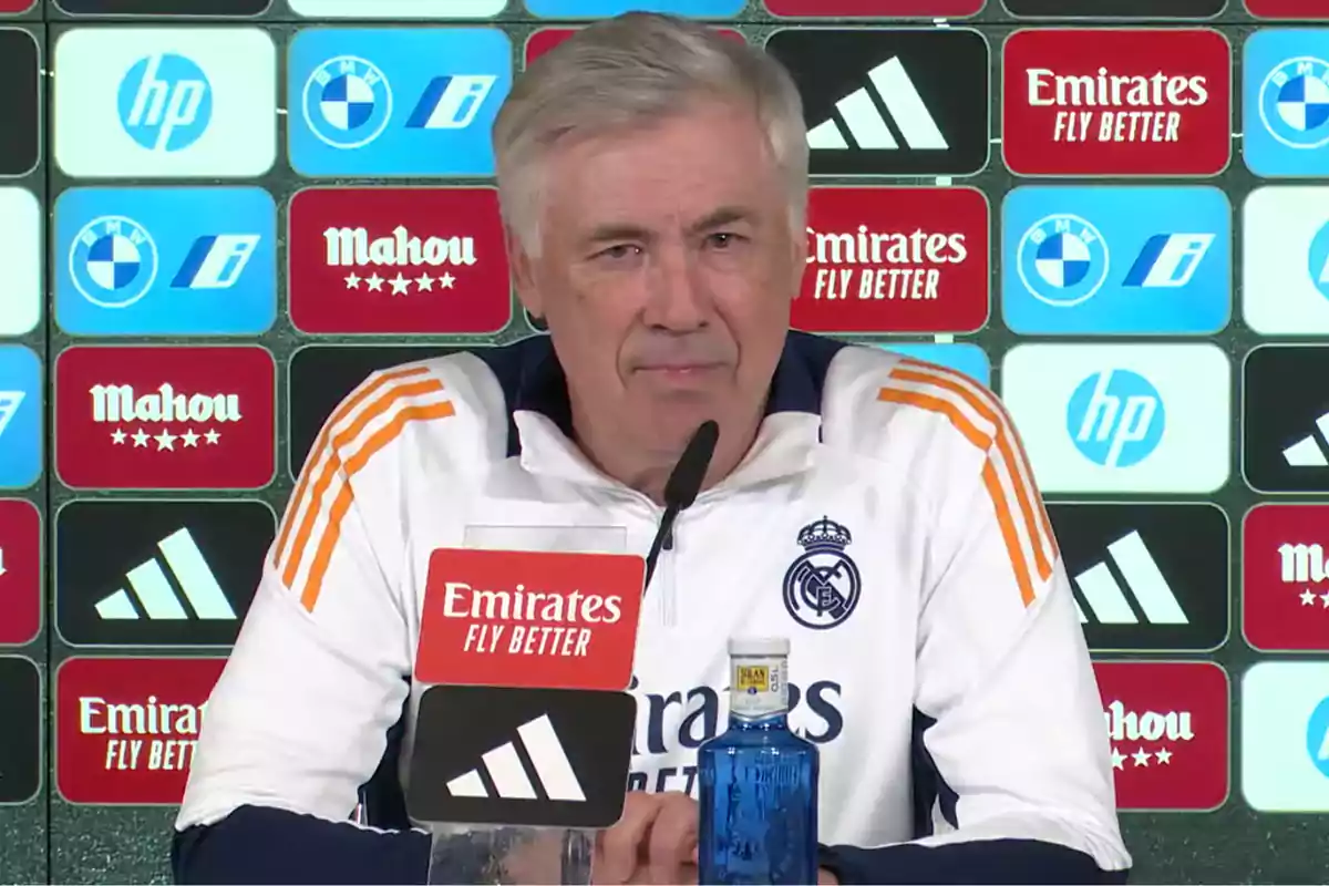 Un hombre en una conferencia de prensa con un fondo de logotipos de marcas y una botella de agua sobre la mesa.