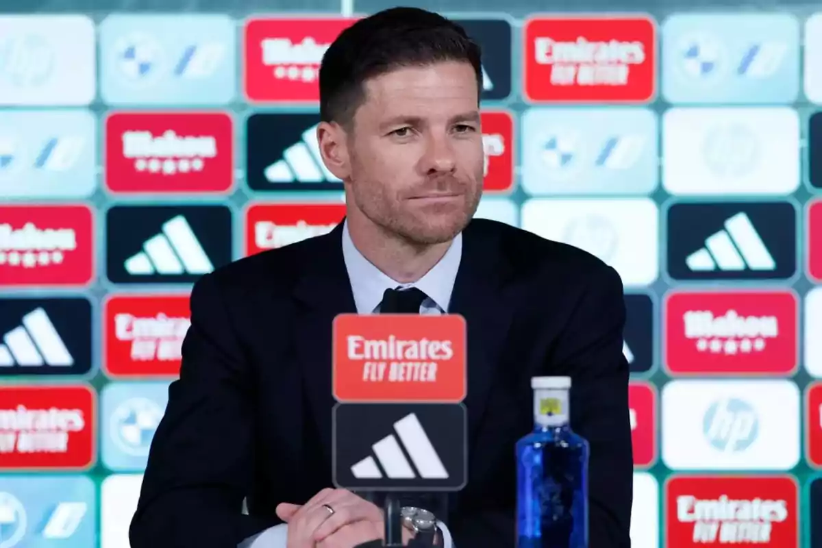 Man in a dark suit sitting at a press conference with a sponsor backdrop and a microphone in front of him Man in a dark suit sitting at a press conference with a sponsor backdrop and a microphone in front of him