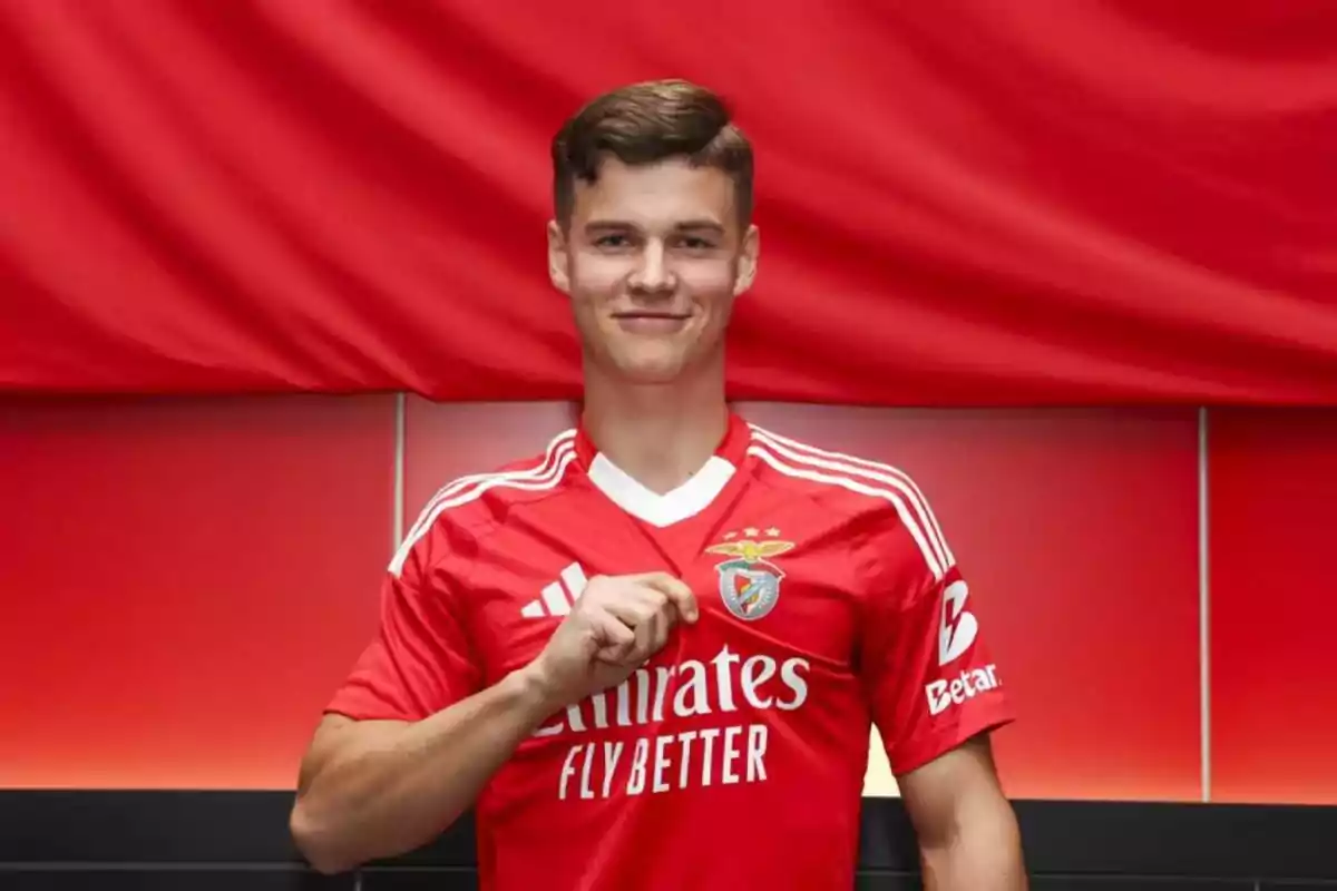 Joven con camiseta roja del Benfica sonriendo y señalando el escudo del equipo frente a un fondo rojo Joven con camiseta roja del Benfica sonriendo y señalando el escudo del equipo frente a un fondo rojo