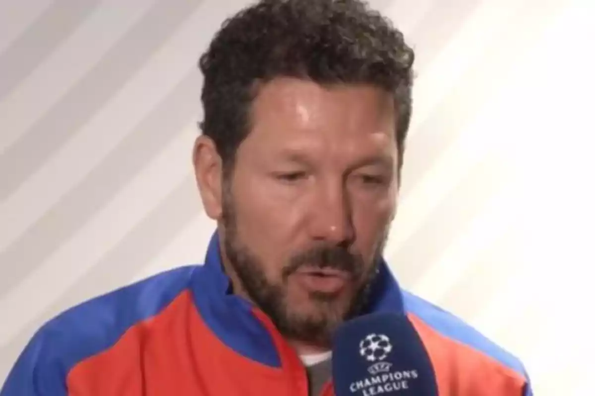 A man with a beard and curly hair is speaking in front of a UEFA Champions League microphone, wearing a blue and red sports jacket.