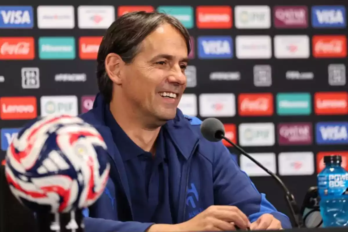 Hombre sonriente con chaqueta azul hablando en una conferencia de prensa con un balón de fútbol y una botella de bebida deportiva sobre la mesa