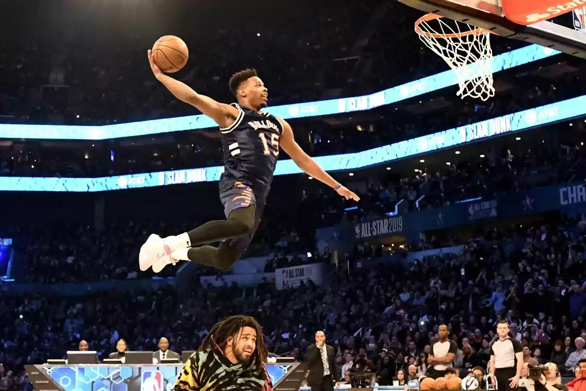 Basketball player performing a spectacular dunk in a slam dunk contest during a sporting event with the audience watching in the background. Basketball player performing a spectacular dunk in a slam dunk contest during a sporting event with the audience watching in the background.