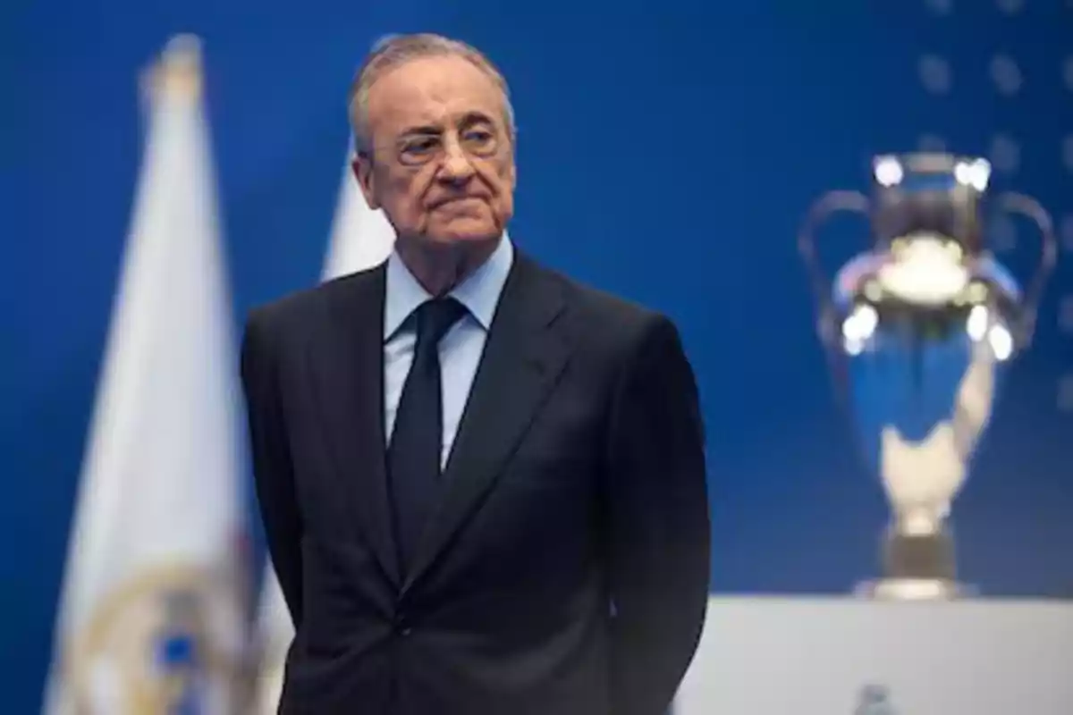 A man in a dark suit is standing in front of a blue background with a trophy cup in the background.