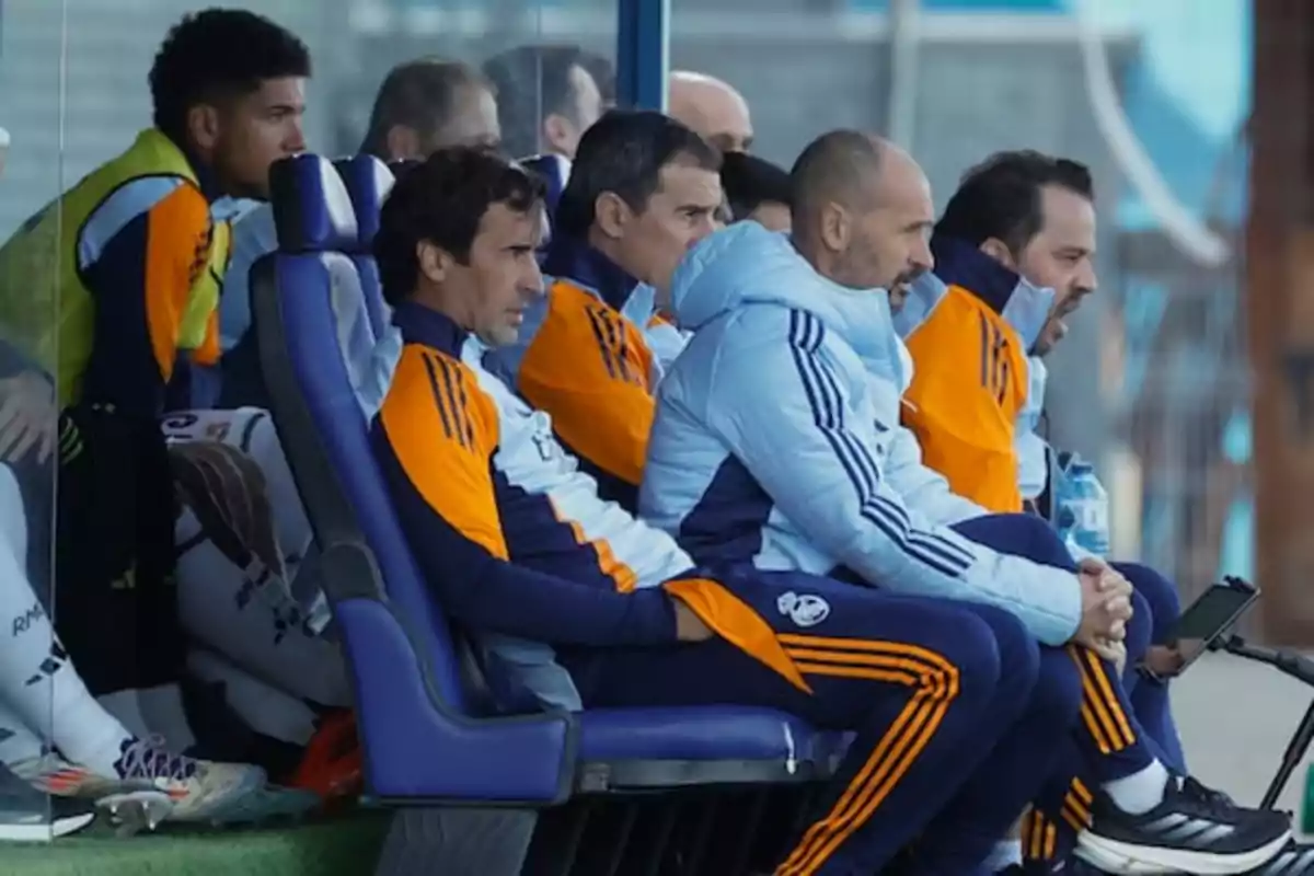 Un grupo de entrenadores y jugadores de fútbol sentados en el banquillo observando el partido.