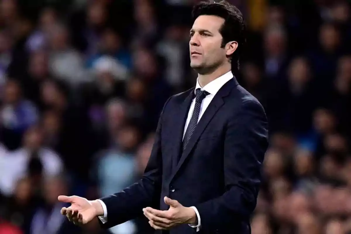 A man in a dark suit with a serious expression and outstretched hands in a stadium setting. A man in a dark suit with a serious expression and outstretched hands in a stadium setting.