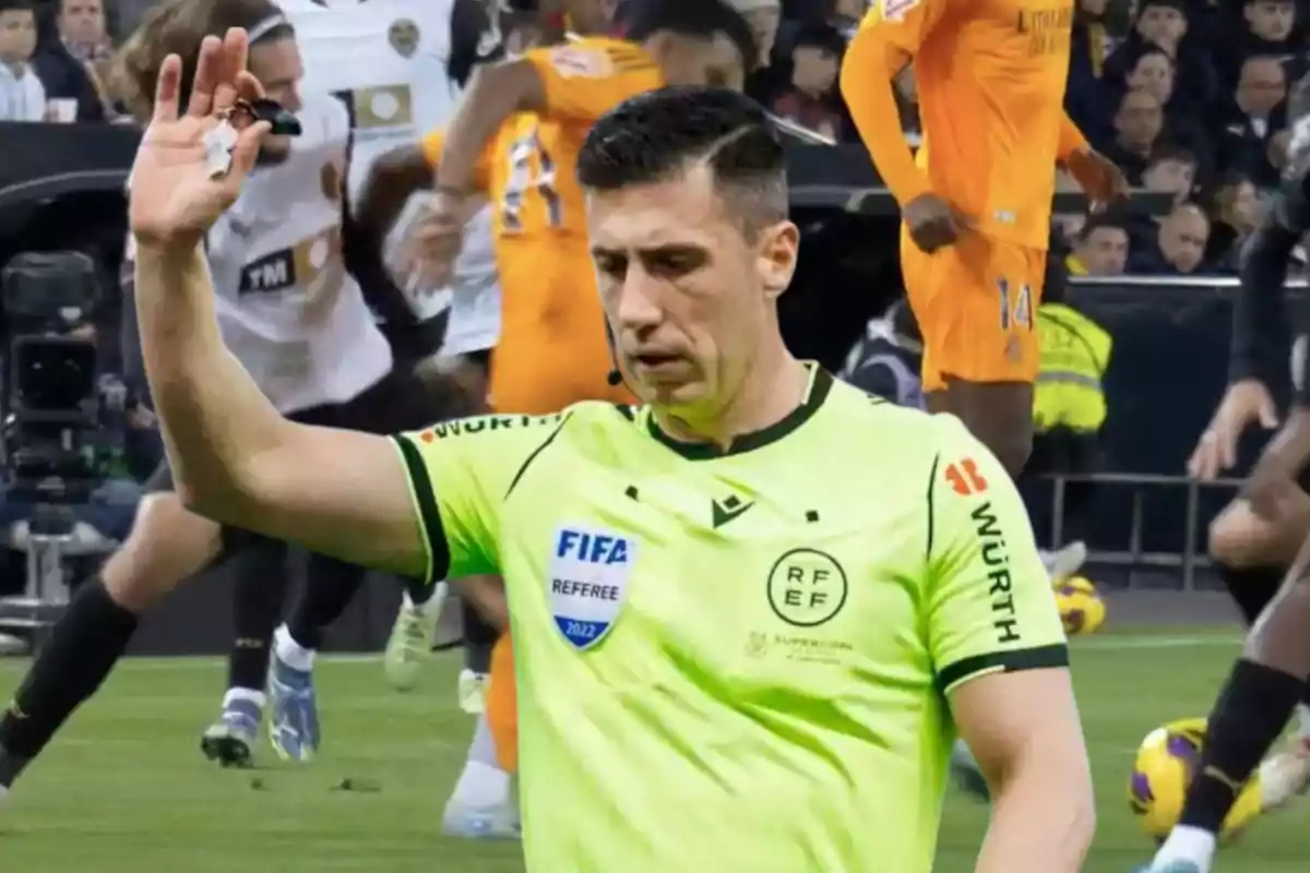 Un árbitro de fútbol en el campo levantando la mano durante un partido con jugadores de fondo.