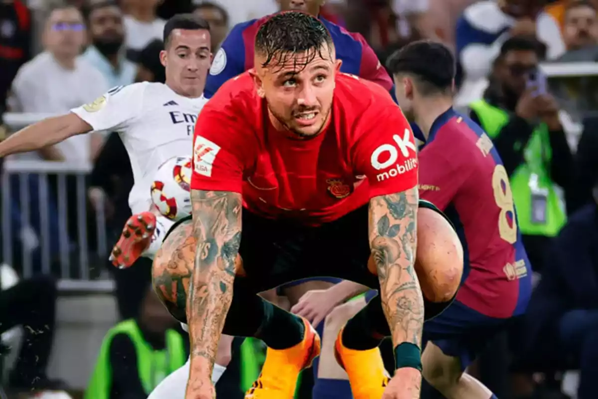 Un jugador de fútbol con tatuajes en los brazos y camiseta roja se agacha en el campo durante un partido, mientras otros jugadores y espectadores se ven al fondo.