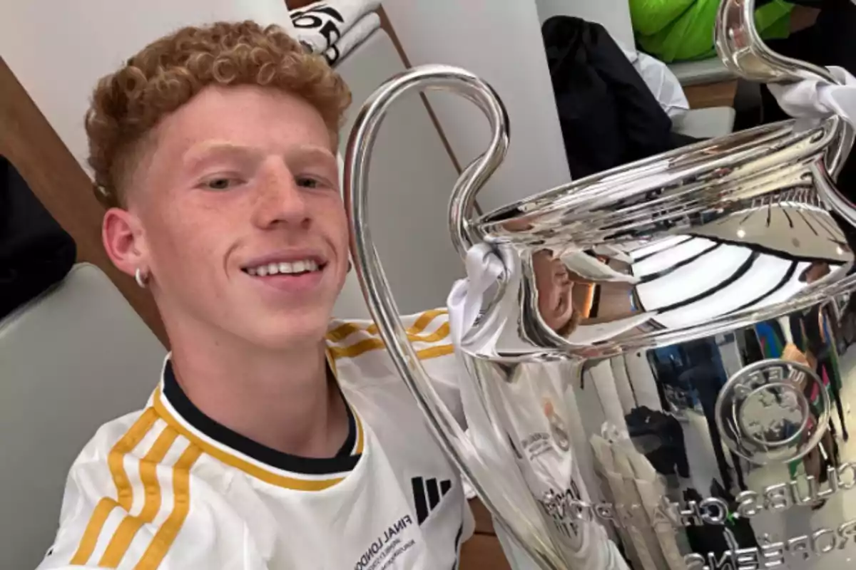 Young man in a white soccer jersey smiles while holding a large silver trophy in a locker room Young man in a white soccer jersey smiles while holding a large silver trophy in a locker room