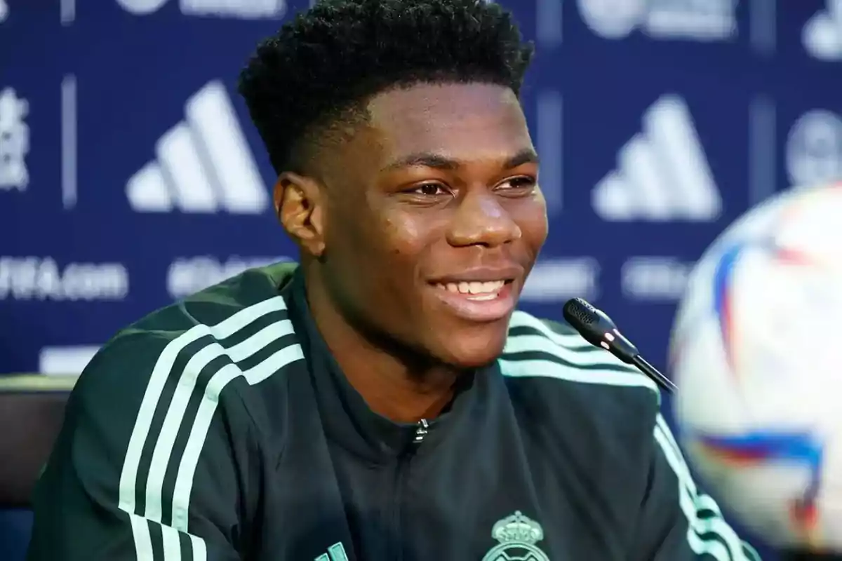 A smiling man in a sports jacket speaks in front of a microphone at a press conference.