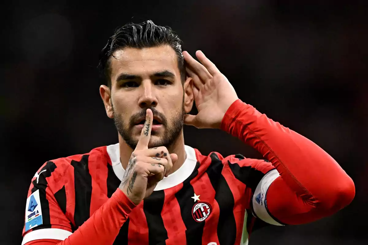 Un jugador de fútbol con uniforme rojo y negro hace un gesto de silencio con el dedo en los labios mientras se lleva la otra mano a la oreja.