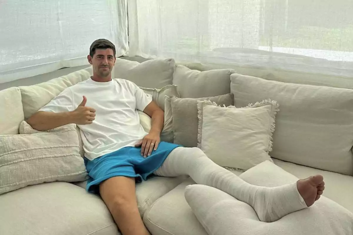 Hombre con pierna enyesada sentado en un sofá con cojines, levantando el pulgar y vistiendo camiseta blanca y pantalones cortos azules. Hombre con pierna enyesada sentado en un sofá con cojines, levantando el pulgar y vistiendo camiseta blanca y pantalones cortos azules.