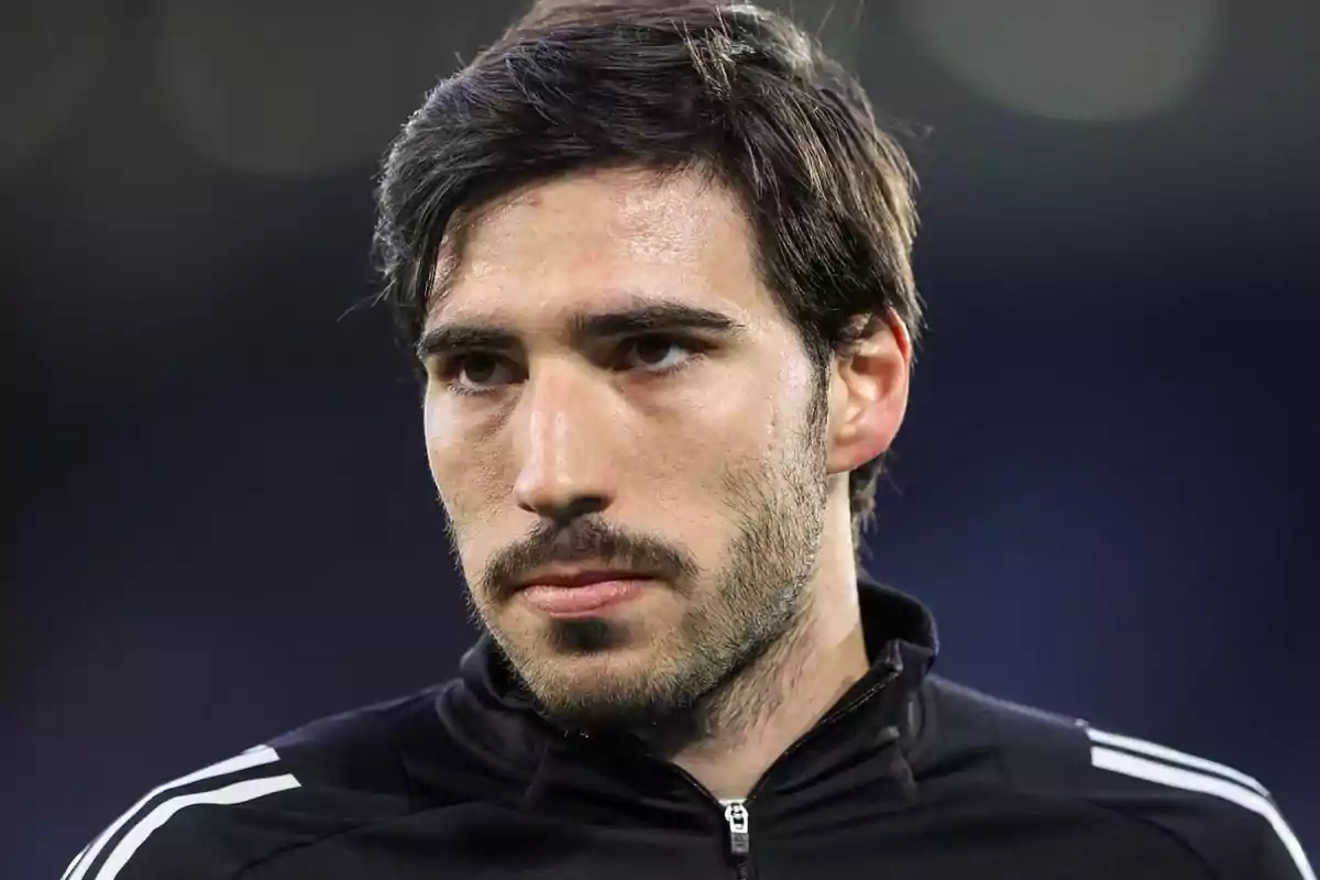 A man with a beard and dark hair is wearing a black sports jacket with white stripes on the shoulders. A man with a beard and dark hair is wearing a black sports jacket with white stripes on the shoulders.