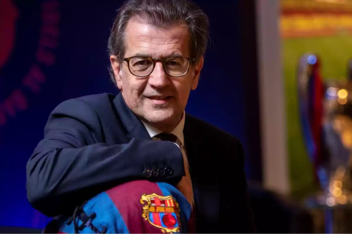 A man with glasses and a dark suit poses with an FC Barcelona scarf.