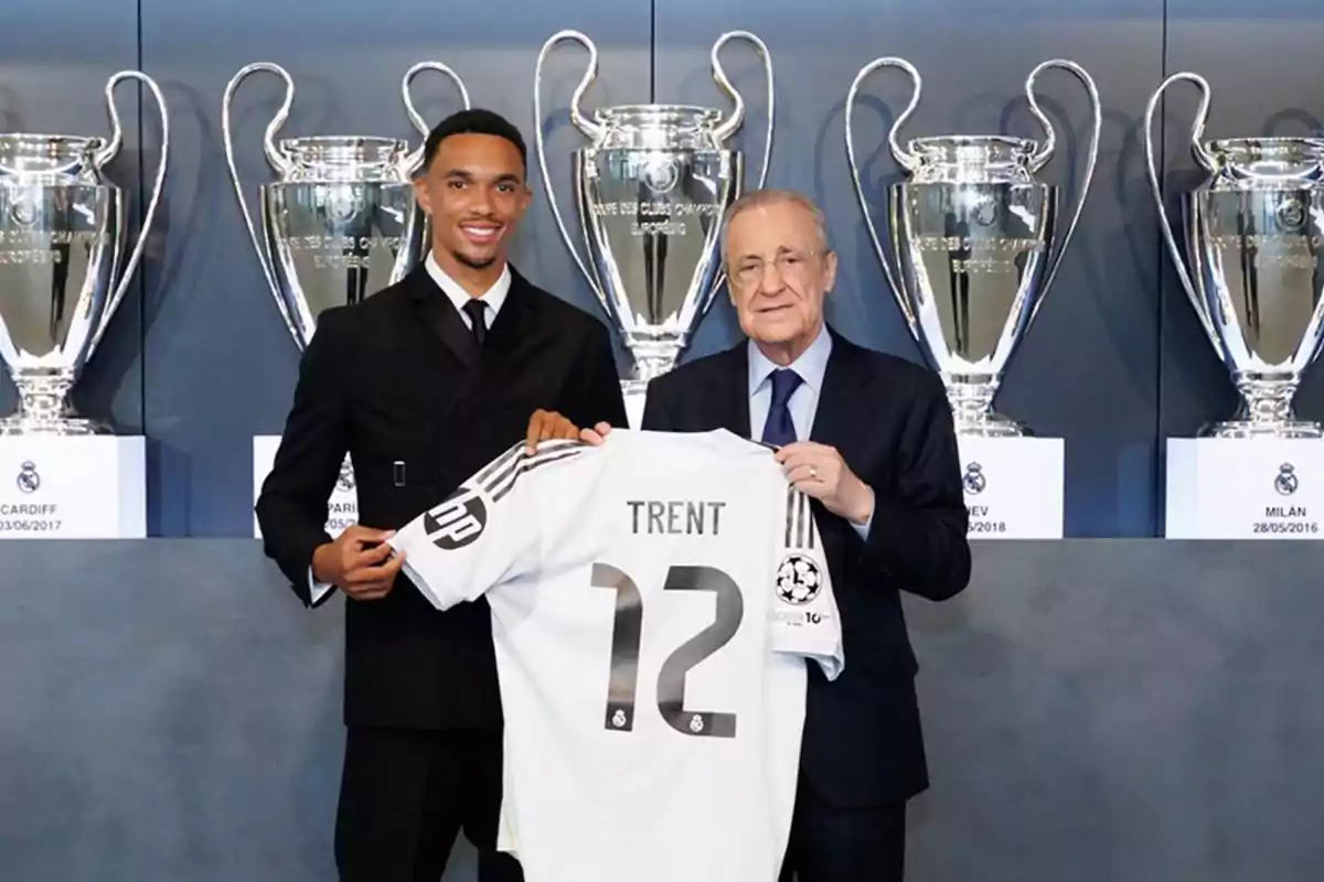 Two people pose smiling while holding a white soccer jersey with the name Trent and the number 12; in the background, there are several Champions League trophies. Two people pose smiling while holding a white soccer jersey with the name Trent and the number 12; in the background, there are several Champions League trophies.
