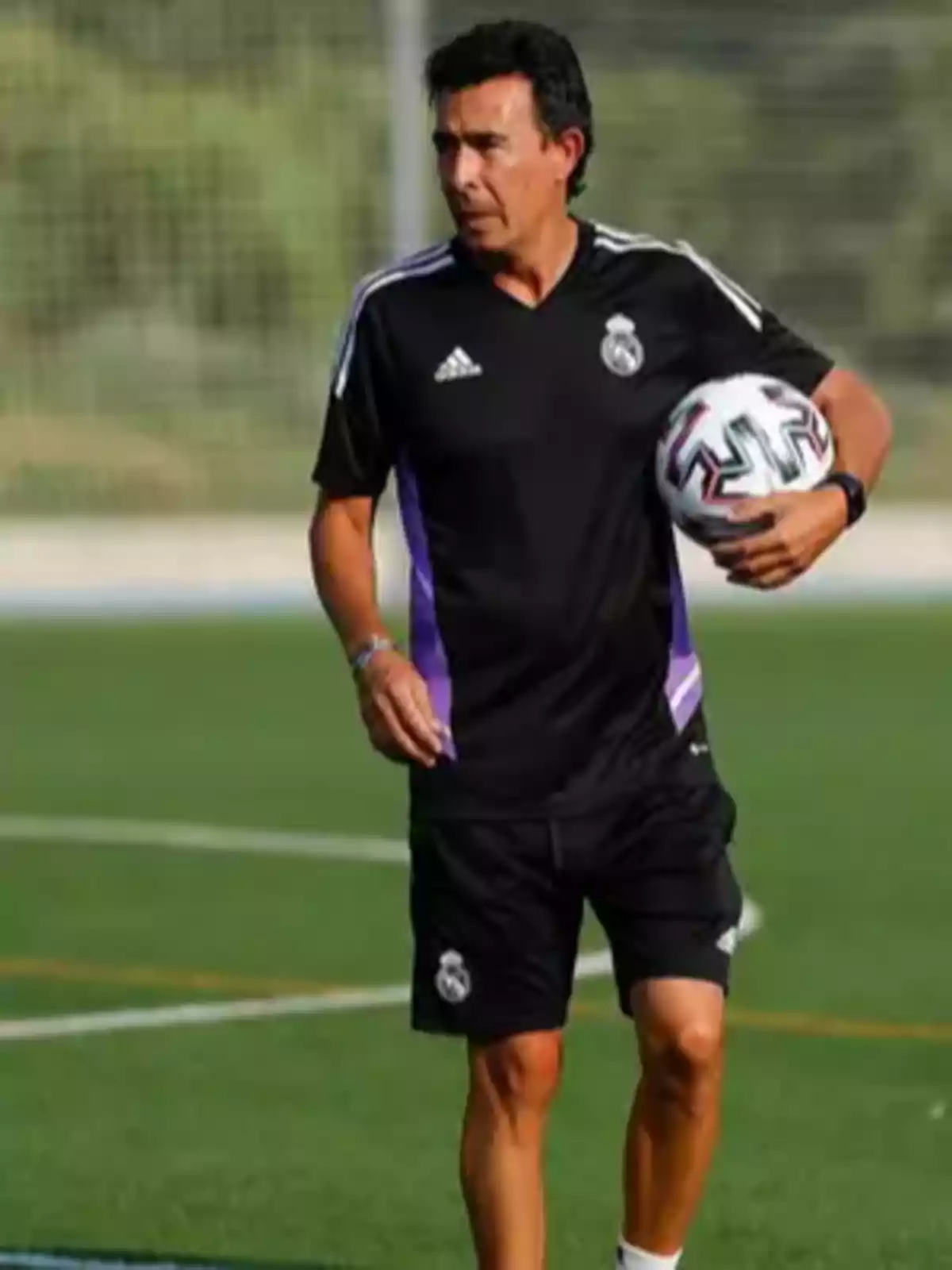 Entrenador de fútbol con uniforme negro del Real Madrid sosteniendo un balón en un campo de entrenamiento