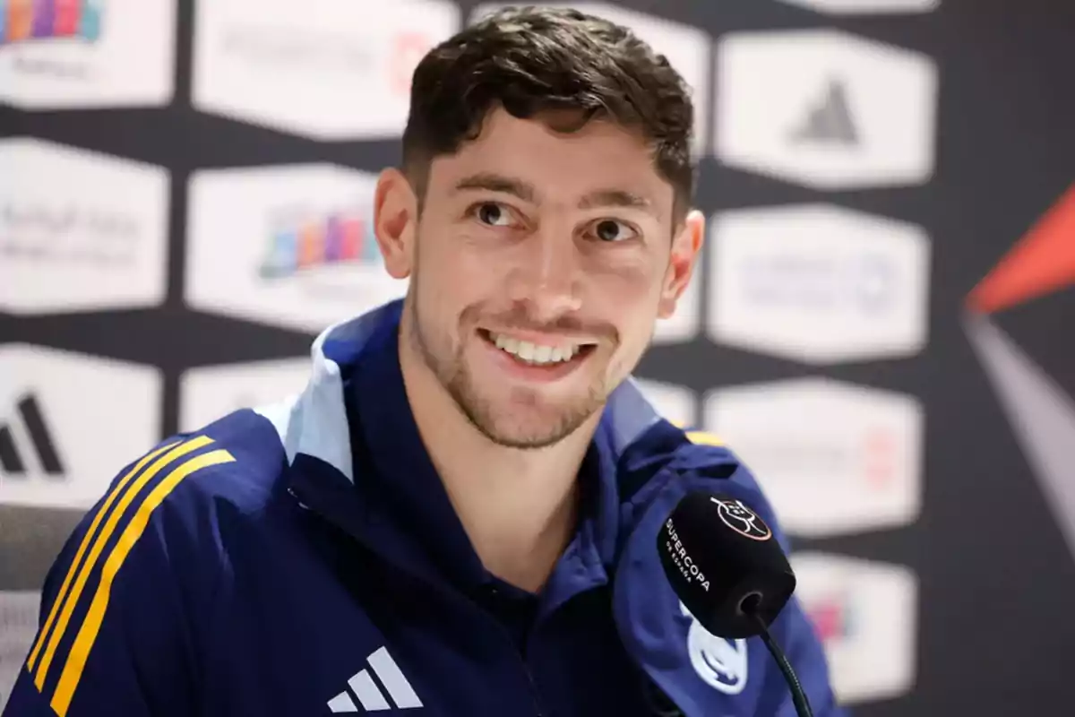 Un hombre sonriente con chaqueta deportiva azul y micrófono en una conferencia de prensa.