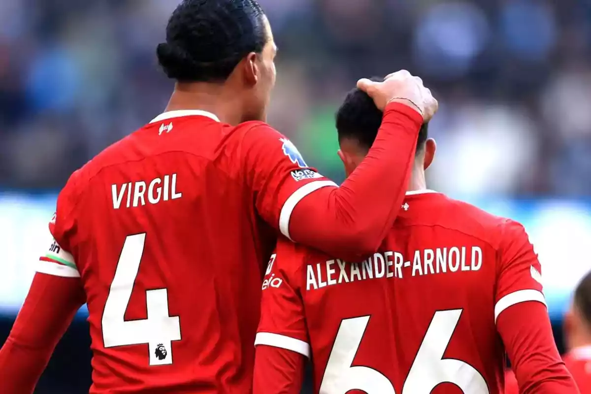 Dos jugadores de fútbol con camisetas rojas, uno con el número 4 y el otro con el número 66, se abrazan en el campo.