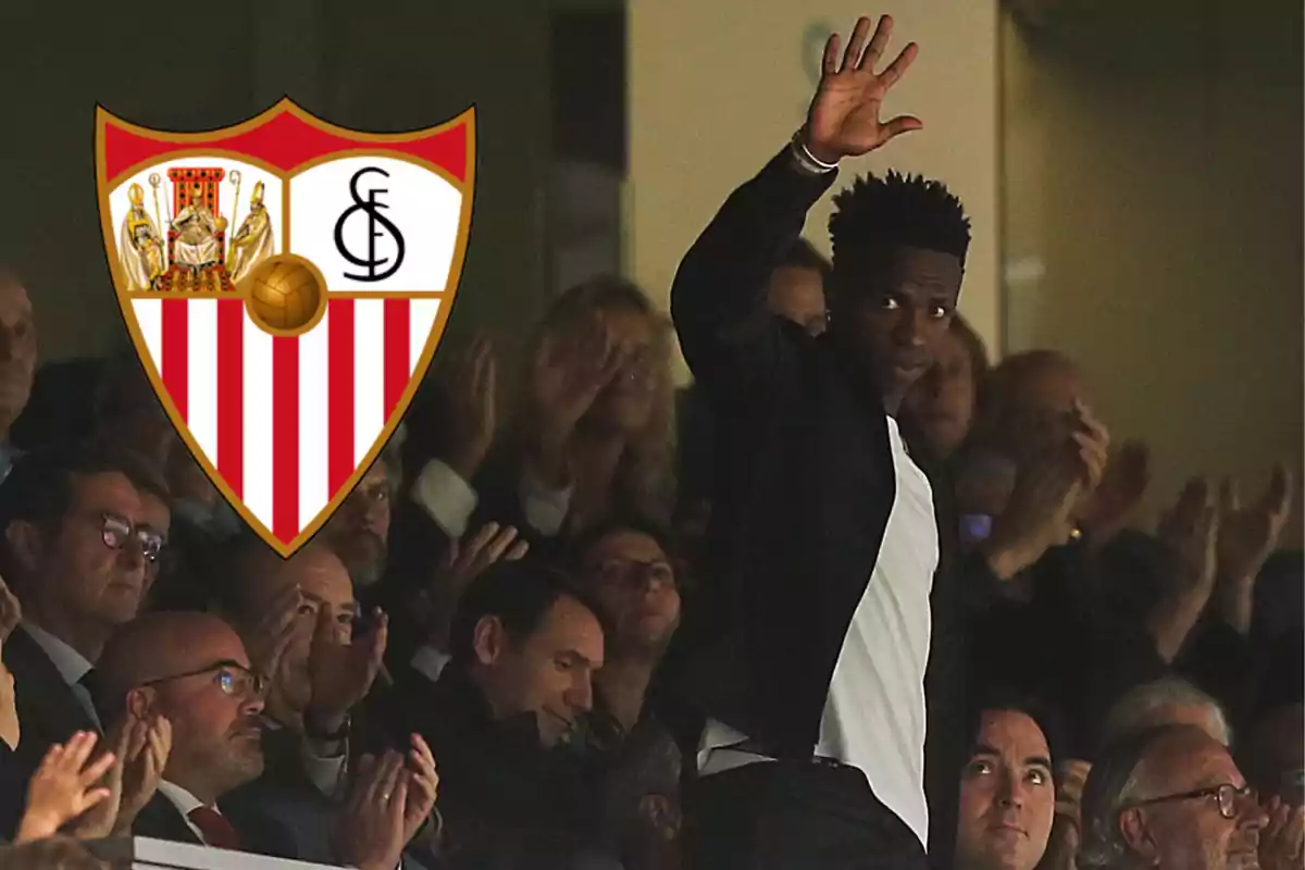 Una persona saluda desde las gradas mientras el público aplaude, con el escudo del Sevilla FC a la izquierda.