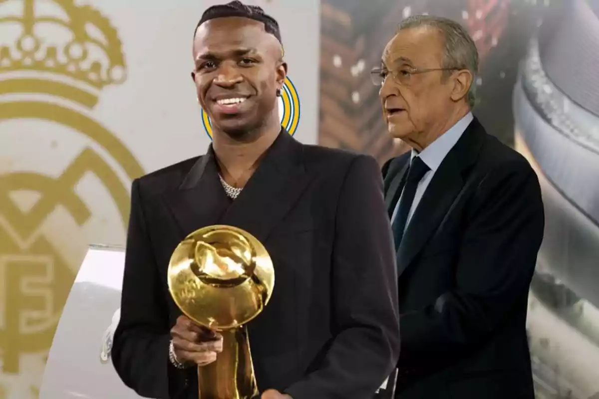 A smiling man holds a golden trophy while another older man stands behind him, both dressed in formal suits.