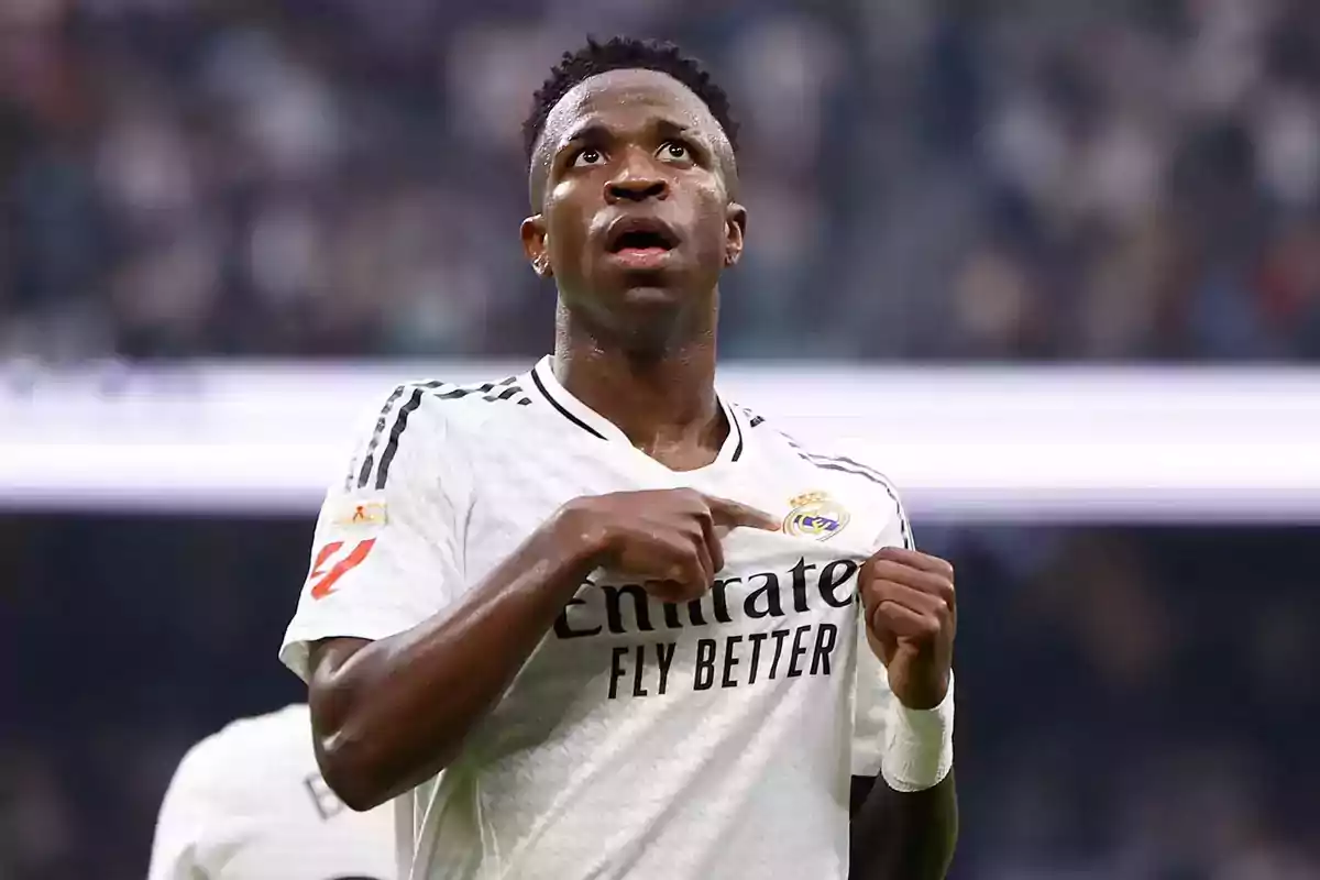 A soccer player in a white uniform points to the crest on his jersey in a stadium.