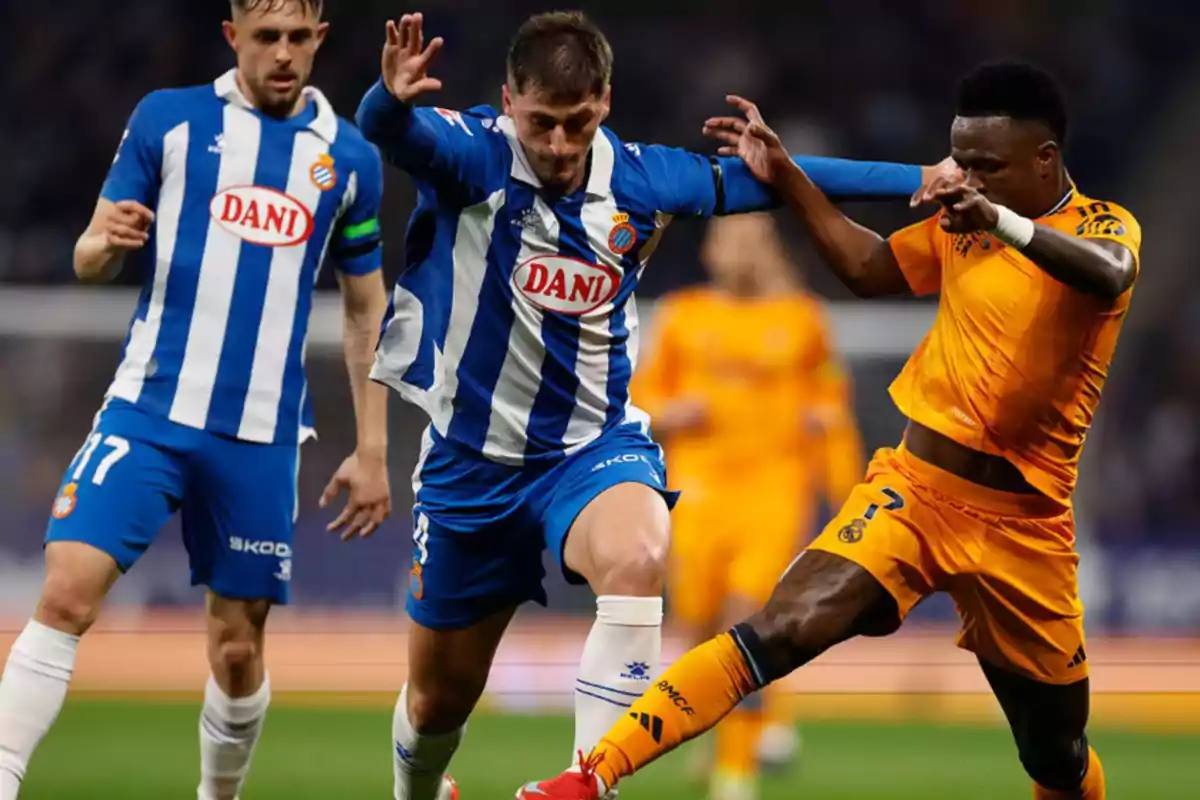 Jugadores de fútbol en acción durante un partido, uno con uniforme azul y blanco y otro con uniforme naranja.