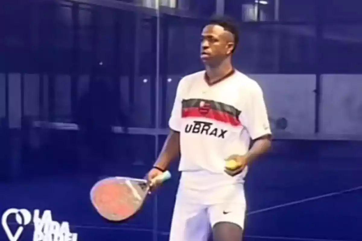 Padel player on a court with racket in hand and ball, wearing a white uniform with red and black details.