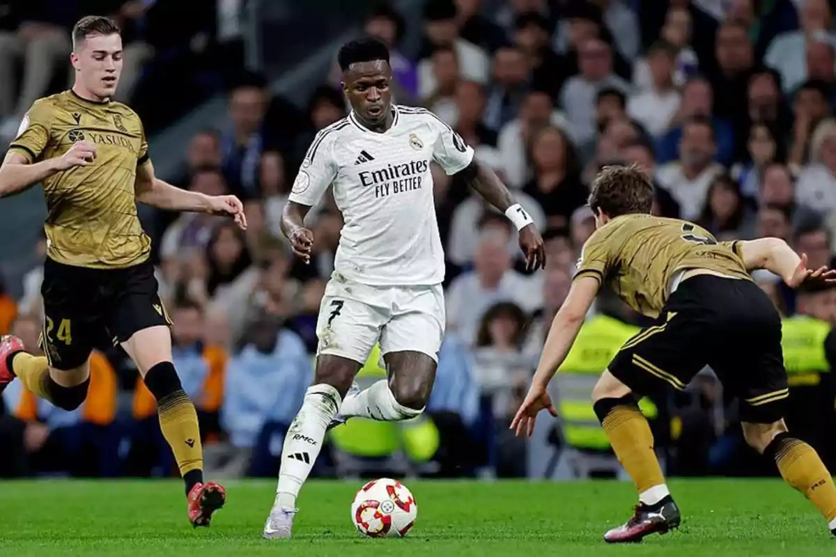 Un jugador de fútbol con uniforme blanco avanza con el balón mientras es perseguido por dos jugadores con uniforme dorado en un estadio lleno de espectadores.