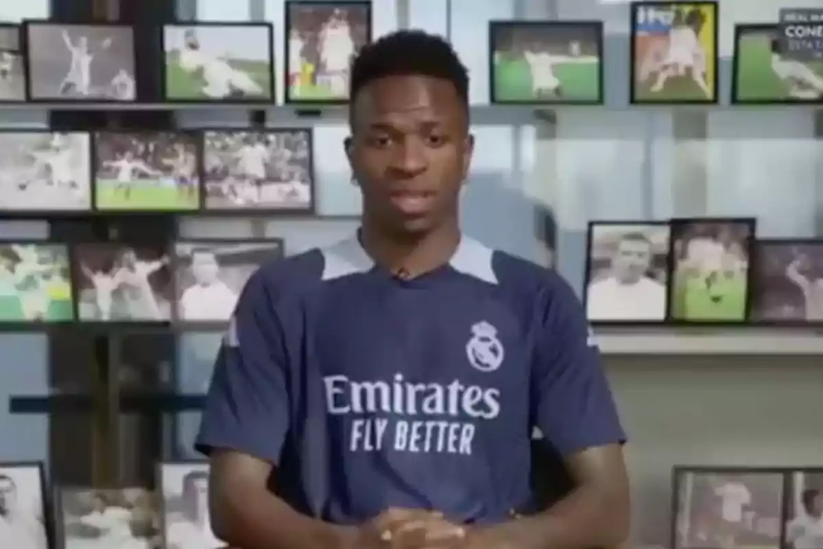 A player wearing a Real Madrid jersey is sitting in front of a wall full of framed photos.