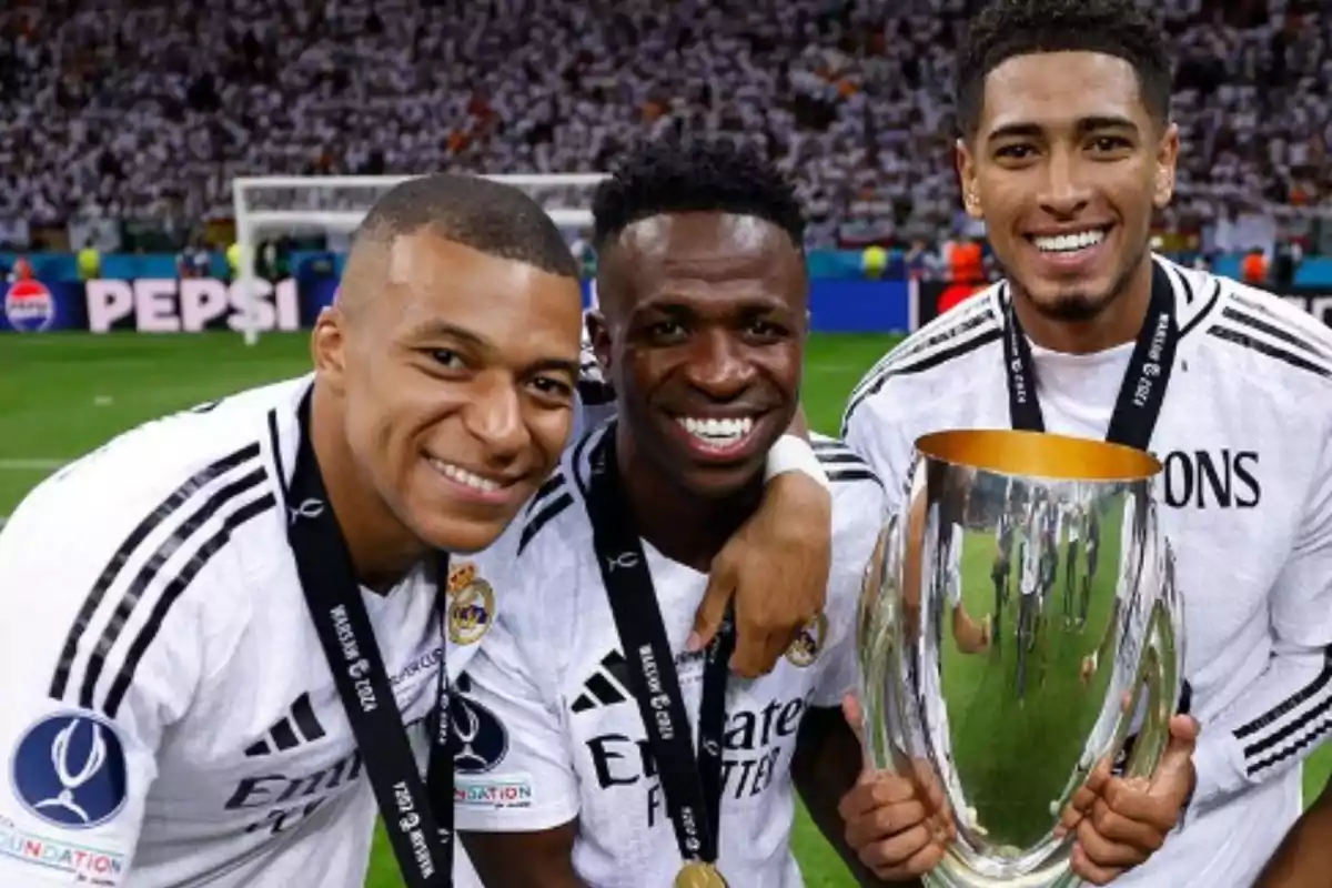 Three soccer players smile as they hold a trophy on the playing field.