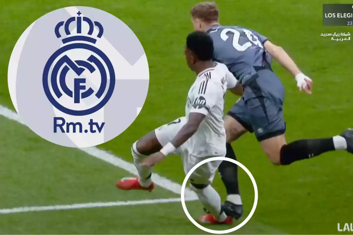 Un jugador de fútbol con uniforme blanco es derribado por un oponente con uniforme oscuro en el campo, mientras el logo de Rm.tv aparece en la esquina superior izquierda.