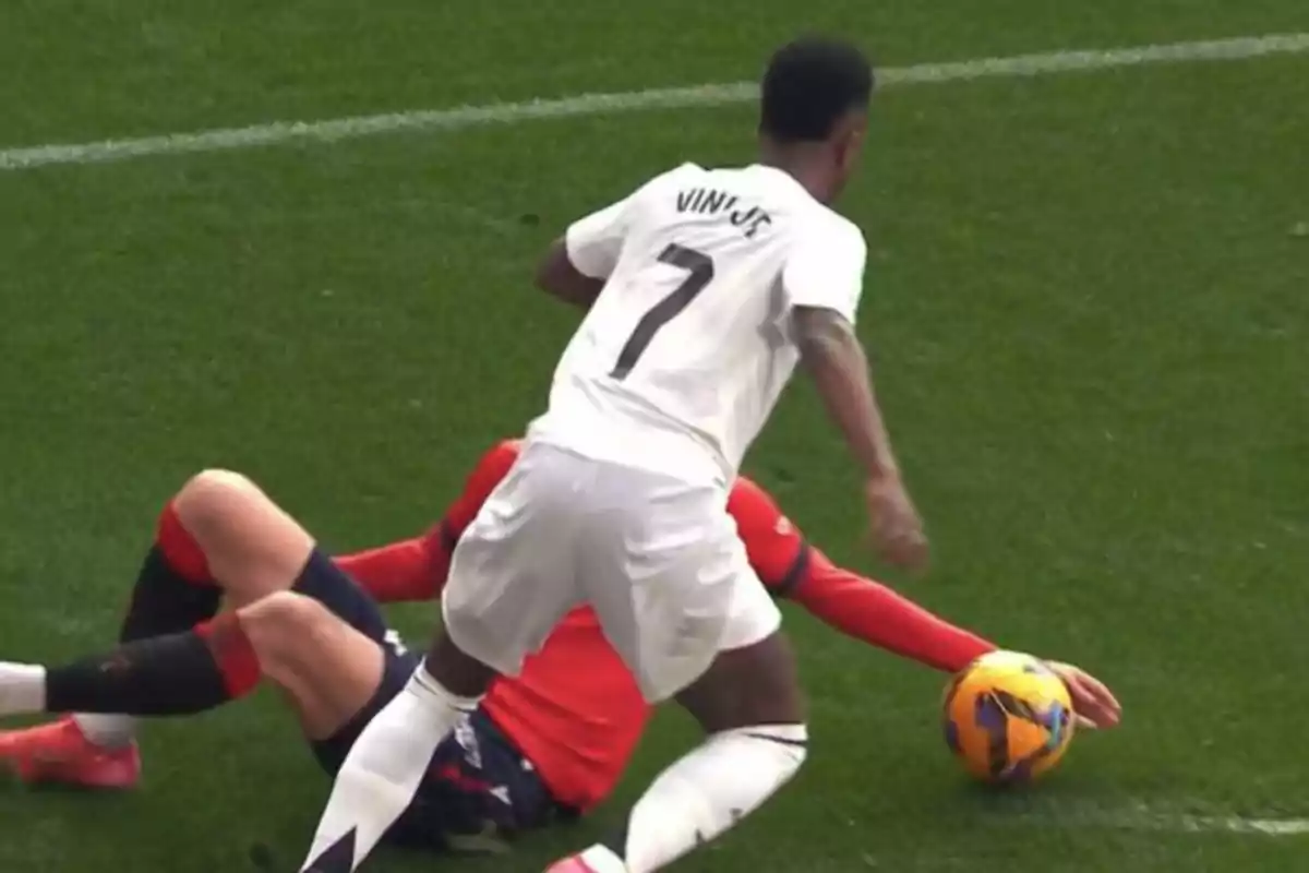 A player with the number 7 on his white jersey dribbles the ball while an opponent in a red uniform tries to stop him on the field.