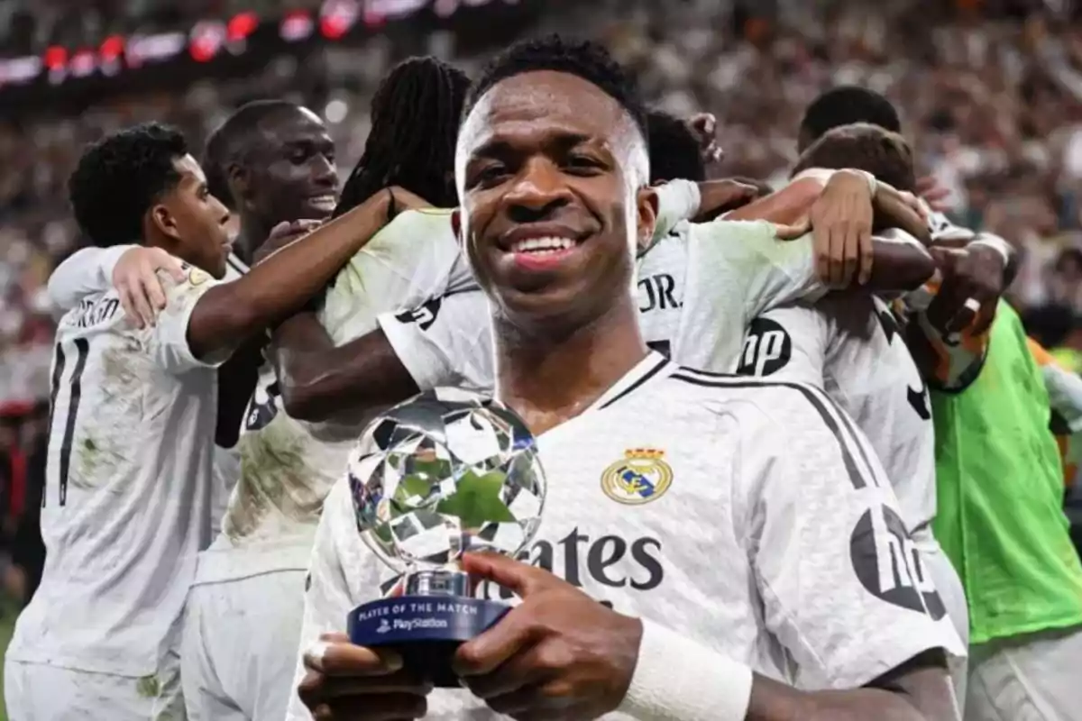 Un jugador de fútbol sonríe mientras sostiene un trofeo, con sus compañeros celebrando en el fondo.