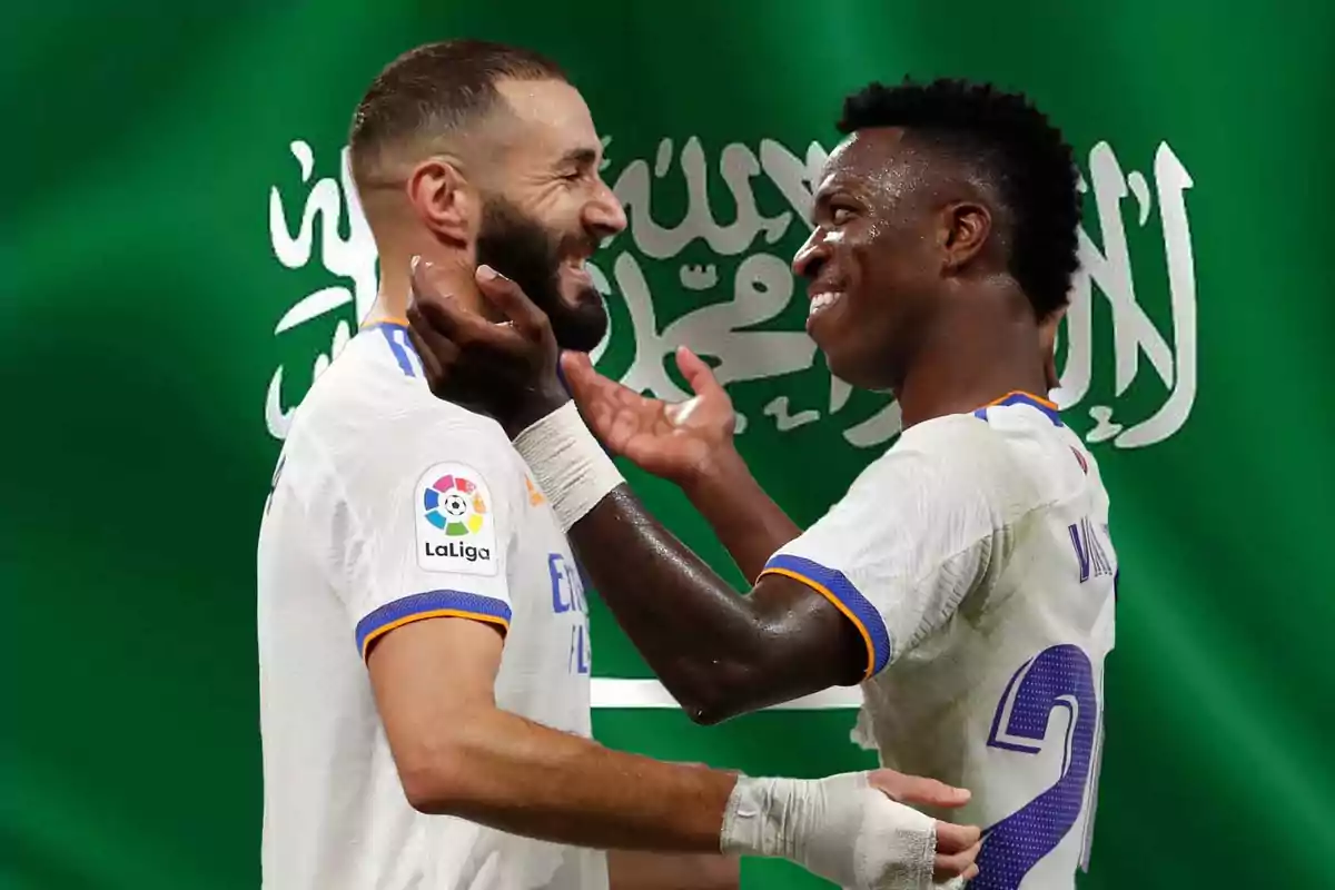 Dos jugadores de fútbol del Real Madrid celebran frente a una bandera verde con escritura árabe.